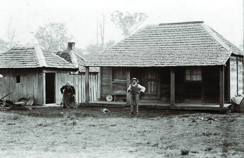 Elderly pioneer settlers outside their timber slab hut, misty morning light